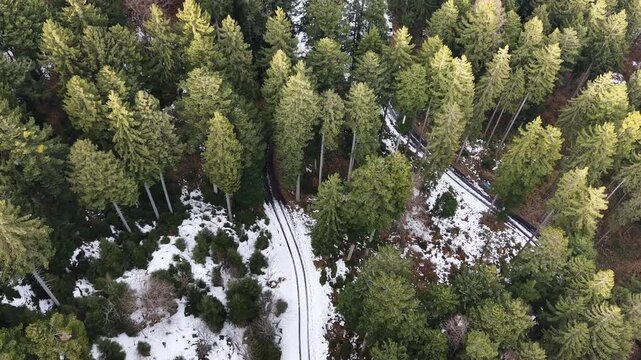 Drone flight along a narrow mountain road after a sharp curve, showing patchy snow on the upper section and clear asphalt below, surrounded by evergreen and leafless trees in winter.