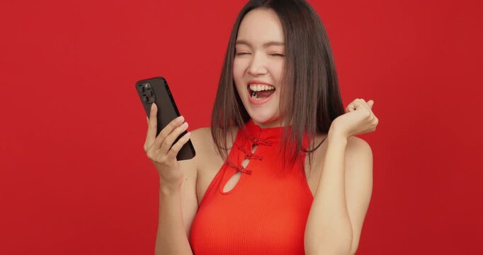 Happy Asian woman in red Chinese dress holding smartphone and celebrating success on red background.