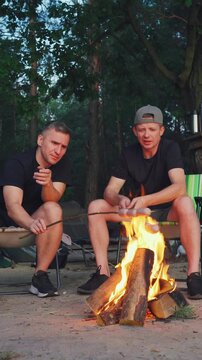 vertical video. Laughter and stories beside glowing campfire, men enjoying friendship and stories in woodland scene, relaxed group sharing humor and tales as night falls in peaceful forest setting