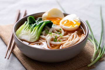 Spicy Udon Noodle Soup topped with Bok Choy, Enoki Mushrooms and Soft Boiled Egg in a bowl with chopsticks on a fabric background