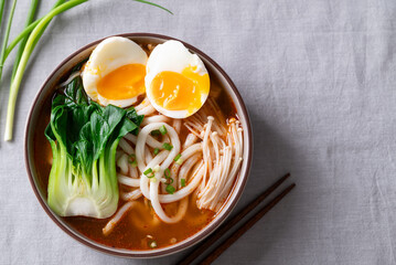 Top view of Spicy Udon Noodle Soup topped with Bok Choy, Enoki Mushrooms and Soft Boiled Egg in a bowl with chopsticks on a fabric background