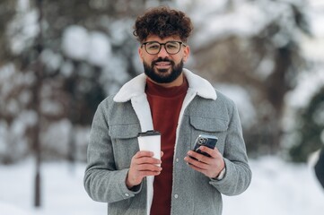 Fototapeta premium Bearded man with glasses wearing a gray jacket and red sweater is holding a coffee cup and smartphone while standing in a snowy outdoor setting with trees in the background