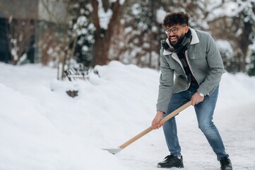Naklejka premium Man with curly hair wearing a gray jacket and blue jeans is shoveling snow in a winter landscape with trees and a path visible in the background during a snowy day