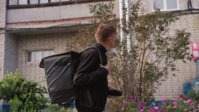 white courier with delivery backpack approaching apartment building adjusting straps and checking smartphone for address and order details on sidewalk near greenery casual jacket focused expression