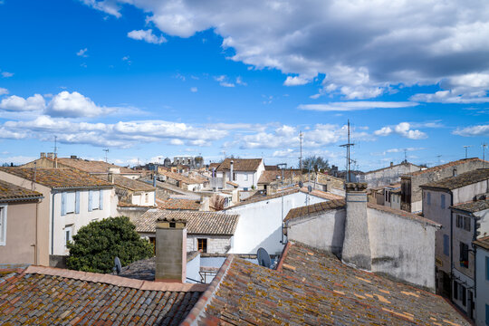 Elevated perspective over traditional tiled rooftops and rustic facades in the historic center of Aigues-Mortes, with dramatic clouds and bright blue sky above. The scene feels airy and textured