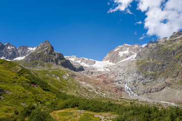 Fototapeta premium Sweeping alpine landscape with Glacier de la Lee Blanche, dramatic rocky peaks, and a striking waterfall cascading down the mountainside under a vivid blue sky with scattered clouds.