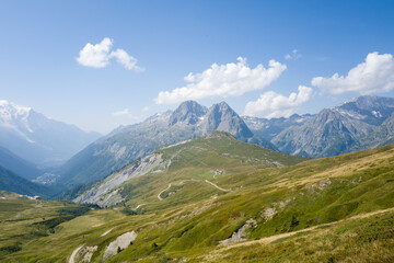 Obraz premium Elevated view of rugged mountain ridges and rolling green slopes with winding trails under a bright blue sky and scattered clouds in the Chamonix valley near Mont Blanc.