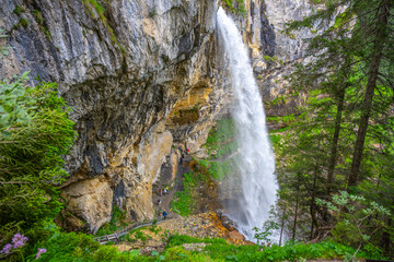 Nestled in the Austrian Alps, Johannes Waterfall cascades dramatically over rocky cliffs, surrounded by vibrant greenery. Visitors explore the area, enjoying the serene atmosphere and stunning views. © pyty