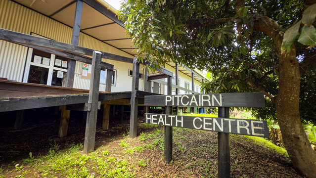 View of the Pitcairn Health Centre sign in front of a long, low wooden building with a dark roof, nestled amongst lush green foliage, Adamstown, Pitcairn Islands.