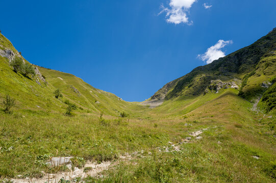 Wide view of a green alpine pass with rugged slopes and scattered rocks, bathed in bright sunlight beneath a vivid blue summer sky near Col du Tricot.