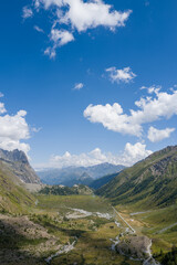Obraz premium Expansive view of a green Italian alpine valley featuring a winding river, rocky slopes, and dramatic peaks beneath a vibrant blue sky scattered with fluffy clouds. Sunlight enhances the textures of