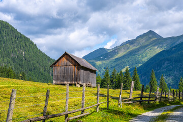 A rustic wooden barn stands beside a winding alpine road in Austria. Surrounded by lush green...