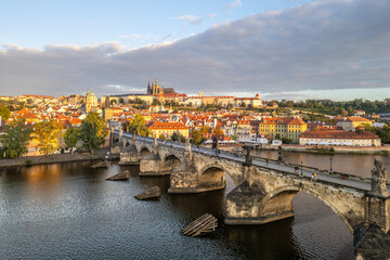 Obraz premium The beauty of Prague Castle rising above the Vltava River, with Charles Bridge in the foreground as the morning sun casts a warm glow over the cityscape.