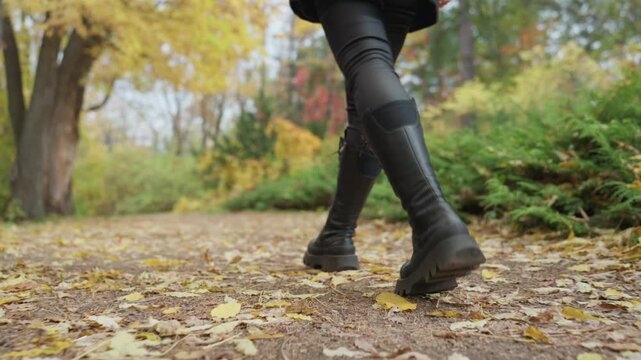 woman in leather boots walking leafstrewn park, closeup legs and boots, confident slow stride, leather pants with knee patches, crunchy fall leaves underfoot, golden foliage and soft bokeh, moody