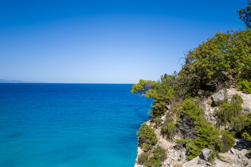 Obraz premium Sunlit Mediterranean vegetation clings to a rocky cliff above the striking turquoise and deep blue sea near Baunei, Sardinia. The clear sky and vibrant coastal colors create a refreshing, tranquil
