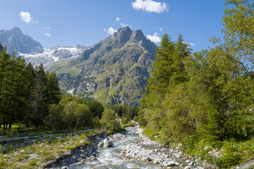 Obraz premium A clear mountain river flows over smooth stones between dense green trees, with rugged peaks and patches of snow rising in the background under a bright blue sky in the Dranse de Ferret valley.