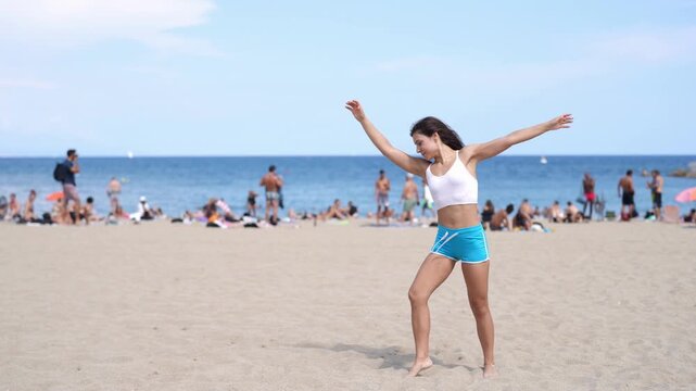 A woman stands on the beach in a blue bikini. She is smiling and she is enjoying herself. The beach is crowded with people, some of whom are also wearing bikinis. The atmosphere is lively and fun