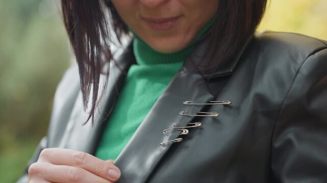 woman fastening safety pin on leather jacket, closeup showing row of metal pins used as brooch, green sweater visible, soft natural bokeh background, autumn light, candid handson styling, edgy DIY
