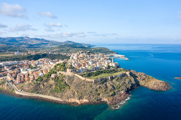 Fototapeta premium Sweeping aerial view of Castelsardo's historic center perched on rugged cliffs, with ancient fortress walls overlooking the vivid blue Mediterranean and rolling Sardinian hills.
