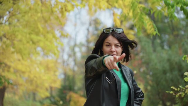 woman in leather jacket walking through autumn park surrounded by yellow leaves and trees, wearing green turtleneck and sunglasses on head, wind blown hair, playful pointing gesture and warm confident