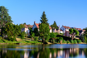 Quaint village homes with red-tiled roofs stand above a vibrant green shoreline, their forms...