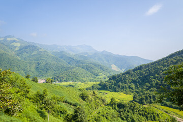 Naklejka premium Sweeping view of terraced rice paddies nestled among green forested hills and distant peaks under a clear blue sky in northern Vietnam. Bright sunlight enhances the vivid natural textures and tranquil