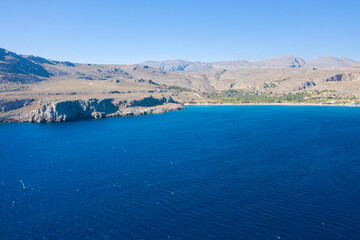 Obraz premium A wide expanse of deep blue water meets sunlit, rugged cliffs and arid hills near Kato Zakros, Crete. The clear sky and textured coastline evoke a sense of openness and natural tranquility.