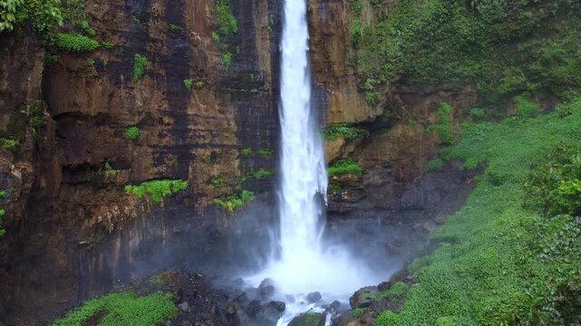 Top view of Koban Kapas Biru Waterfall, East Java. Water cascades down a high cliff into a lush green canyon surrounded by tropical forests. Lumajang River. Dron. 4K