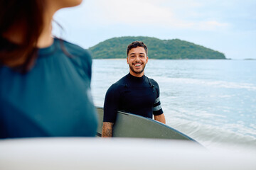 Fototapeta premium Young man wearing a wetsuit and holding a surfboard standing on the beach with the ocean and an island in the background