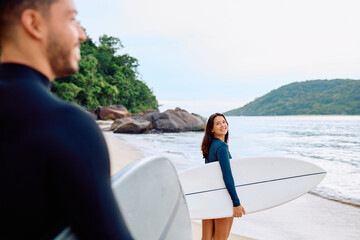 Fototapeta premium Young couple carrying surfboards on tropical beach. Woman standing on sand looking back and smiling. Man standing in foreground