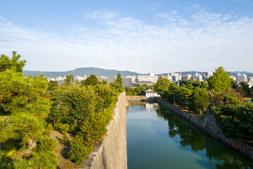 Fototapeta premium Elevated perspective of a tranquil moat lined with historic stone walls and lush greenery, with Kyoto city and distant mountains under a bright blue sky.