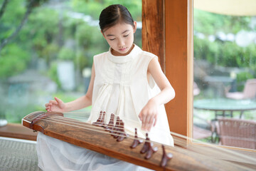 A young girl is playing a traditional Chinese instrument, the guzheng. She is sitting on the floor...