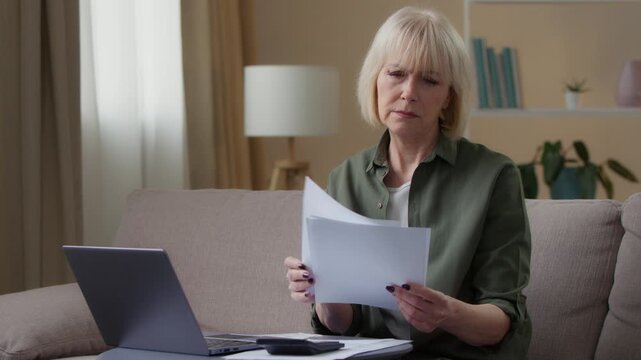 Serious Caucasian old woman with papers documents looking laptop confused senior female retired lady checking data letters income budget expenses with computer at home homeowner paperwork indoors