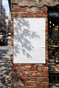 Blank white poster mockup glued to a weathered brick wall on a sunny urban street with tree shadows