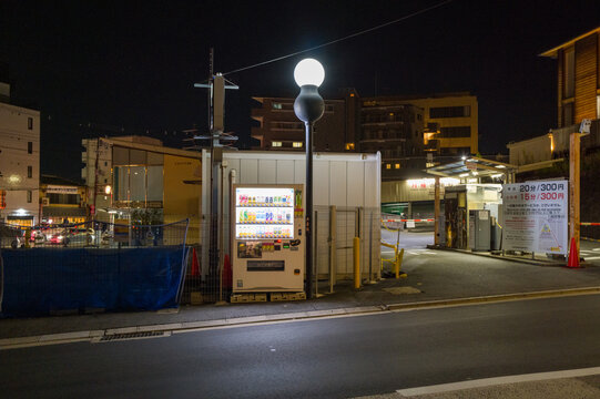 Brightly lit vending machine stands outside a parking lot in Kyoto at night, surrounded by urban buildings and glowing streetlights, creating a quiet city atmosphere with deep shadows and artificial