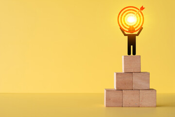 Silhouette of business person standing on wooden block stairs holding glowing target board icon with arrow.Success,goal achievement, leadership,winner concept on yellow background with copyspace.
