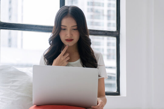 Thoughtful young Asian woman working on laptop computer sitting on bed near window city view. Serious female freelancer or student looking at screen with finger on chin. Remote work online education.