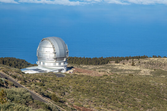 Side view of the Gran Telescopio Canarias dome in La Palma, situated on a high volcanic plateau above the clouds and the Atlantic Ocean, surrounded by native high-altitude vegetation.