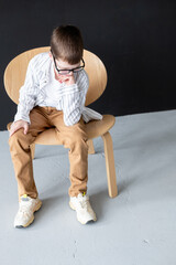 An 11-year-old boy wearing reading glasses and a hearing aid, wearing a white shirt, sits on a chair against a black wall, in a pensive pose