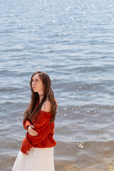 A young woman with long hair and blue eyes, in a white skirt and a terracotta sweater, stands barefoot in the water against the backdrop of the sea