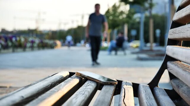 Man bending down to pick up a forgotten phone from wooden park bench