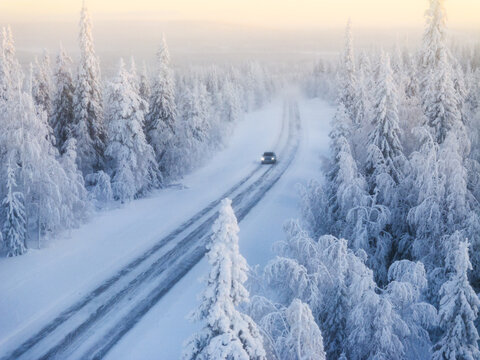 Aerial view of a lone car navigates a snow-laden road cutting through a pristine forest of frosted trees, Lapland, Finland.