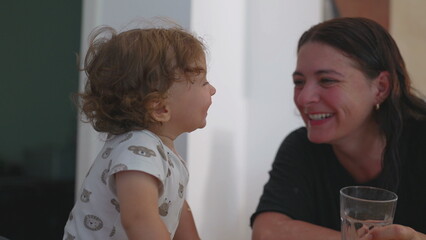 Mother helps toddler drink water from glass while smiling, warm and funny moment as baby sips...