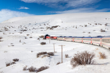 Eastern Express train entering a tunnel in Erzincan, Turkey. Red locomotive and carriages against snowy mountains and winter sky. Iconic Turkish rail journey in the frozen wilderness.