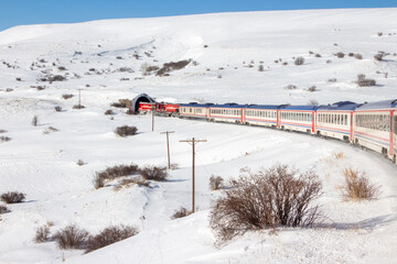 Eastern Express train entering a tunnel in Erzincan, Turkey. Red locomotive and carriages against snowy mountains and winter sky. Iconic Turkish rail journey in the frozen wilderness.