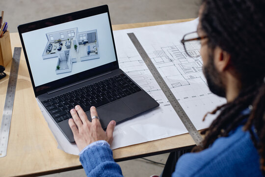 Middle aged Black man working at desk viewing 3D architectural floor plan on laptop, hand resting on keyboard, technical drawings and rulers visible on workspace, focusing on project