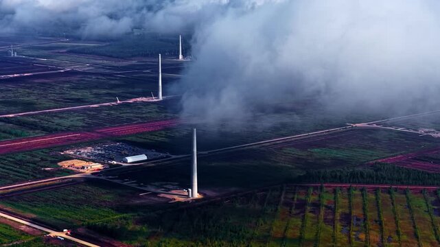 Drone shot of renewable energy project site showing several tall white turbine towers standing in fields and forests of Latvia