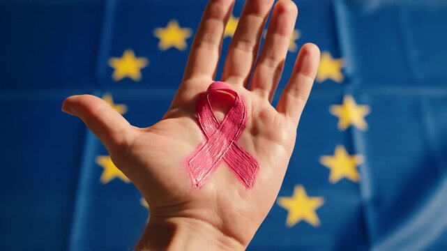 A pink painted ribbon on a hand, set against a blurred flag of Europe in the background