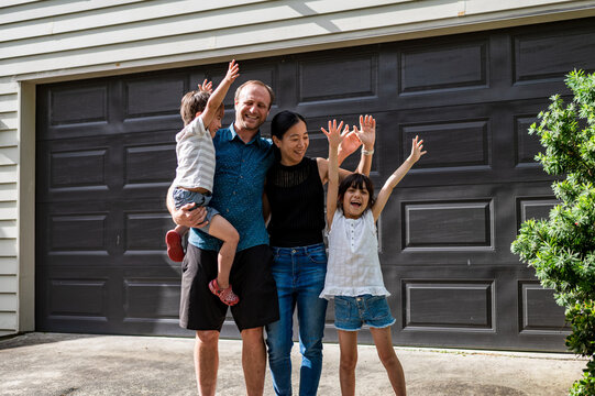 A family of four stands in front of their house, smiling and cheering joyfully on a sunny day.