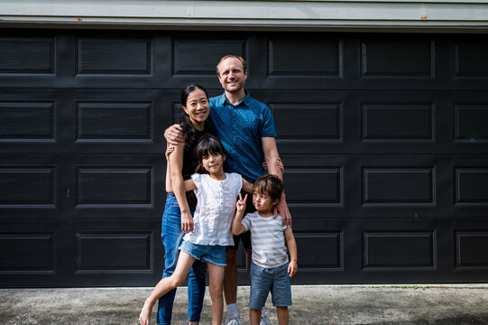 Family of four stands happily together outside their home, smiling in portrait